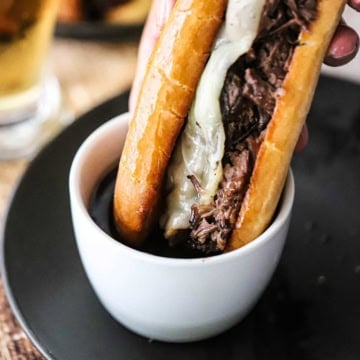 A close-up view of a person plunging a slow cooker French dip sandwich into a small white bowl filled with an au jus sauce sitting on a black dinner plate.