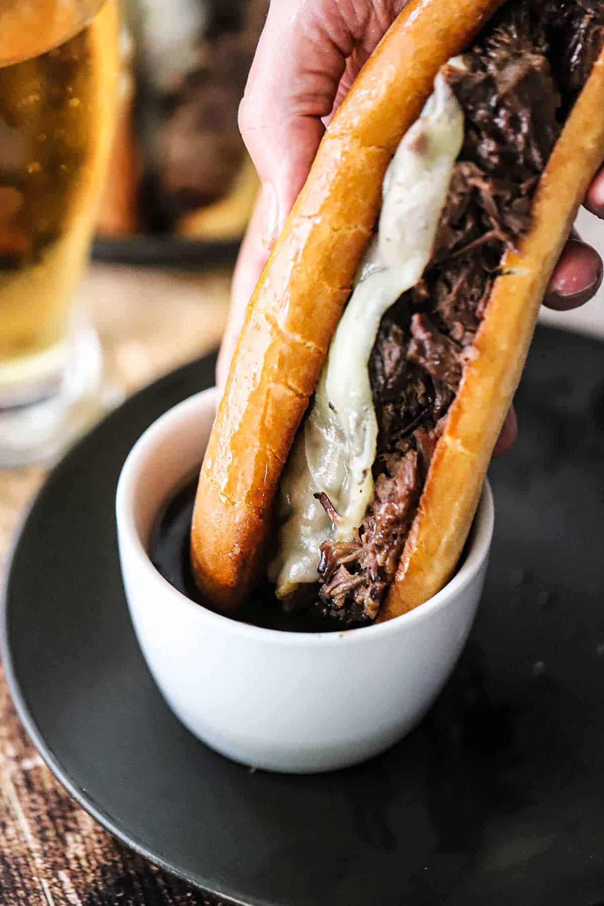 A person plunging a slow cooker French dip sandwich into a small white bowl filled with an au jus sauce sitting on a black dinner plate.