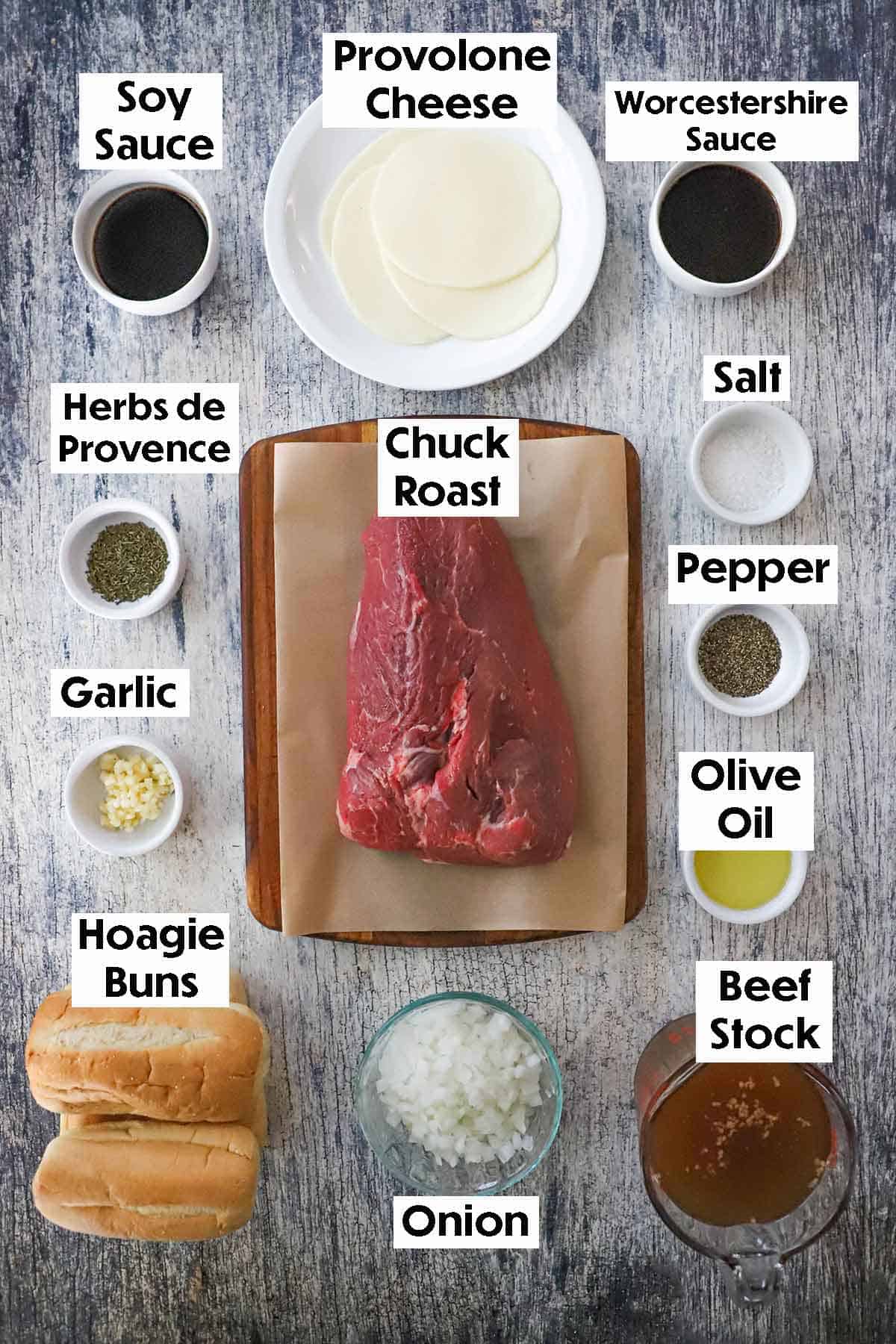 An arrangement of ingredients for slow cooker French dip sandwiches on a grey wooden background including a chuck roast, soy sauce, Worcestershire sauce, beef stock, onion, olive oil, and seasonings.