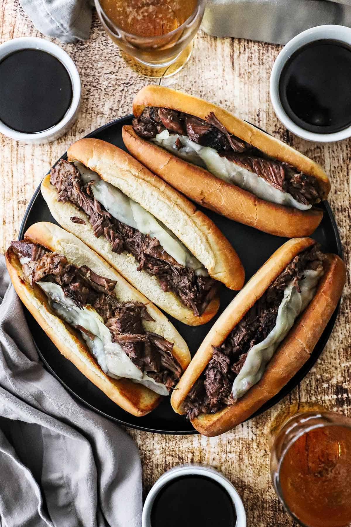 An overhead view of four large slow cooker French dip sandwiches resting on a large black dinner plate with several bowls of au jus sauce nearby.