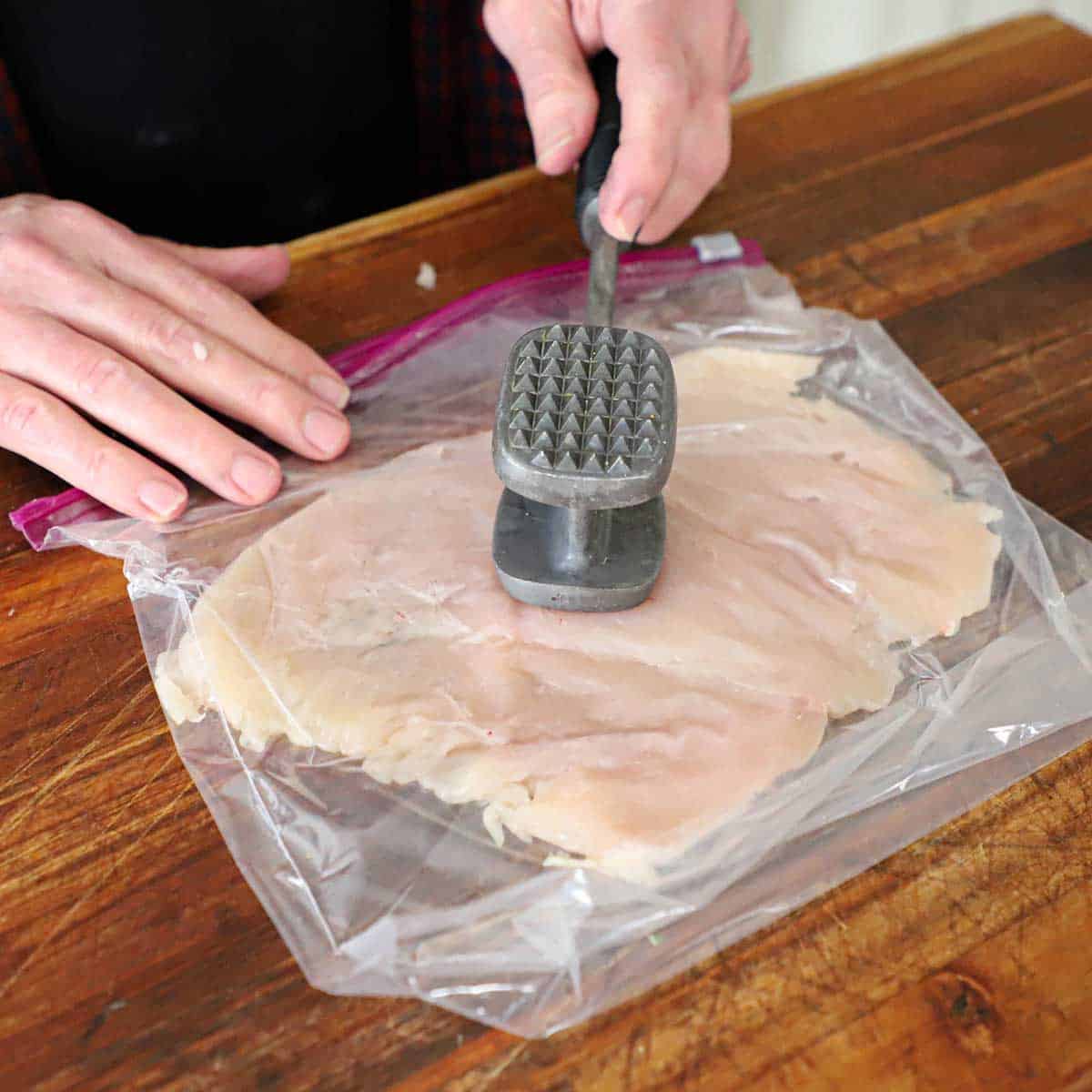 A person using a metal mallet to pound a chicken breast inside a plastic baggie on a wooden cutting board.