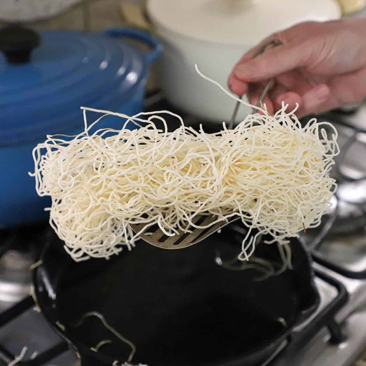 A person holding freshly fried vermicelli rice noodles that have puffed and are a bright white over a cast-iron skillet filled with hot oil on a gas stove.