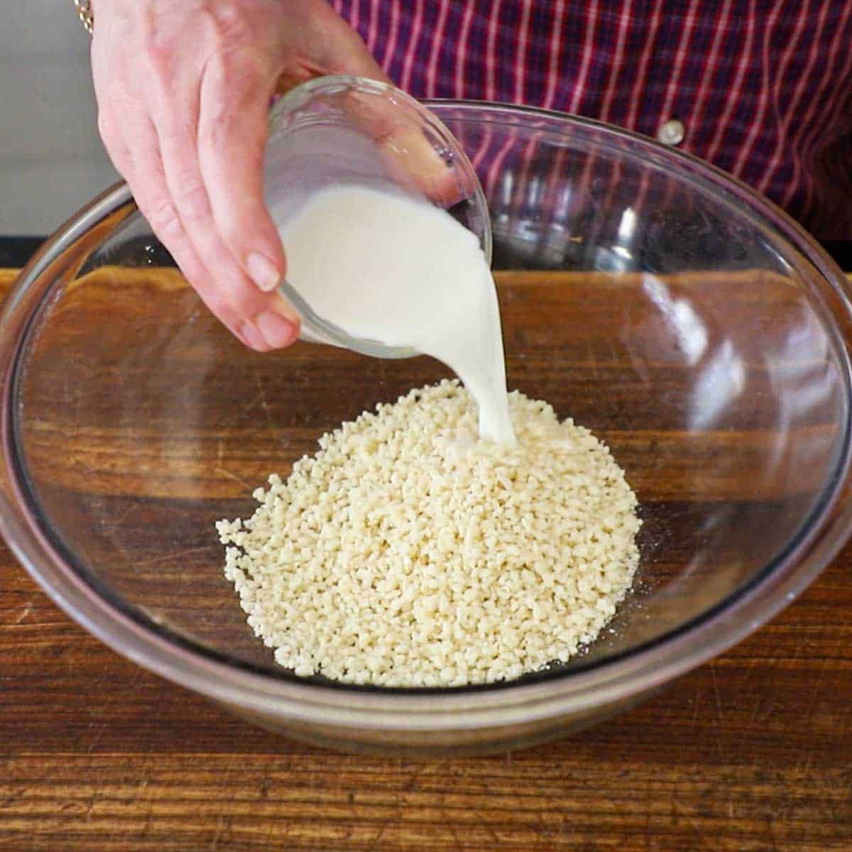 A person pouring whole milk from a small glass bowl into a large bowl filled with 1 cup of Panko breadcrumbs.