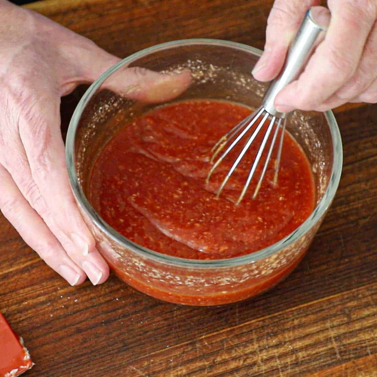 A person using a small whisk to combine ketchup, horseradish sauce, lemon juice, hot sauce, and Worcestershire sauce in glass bow for homemade cocktail sauce.