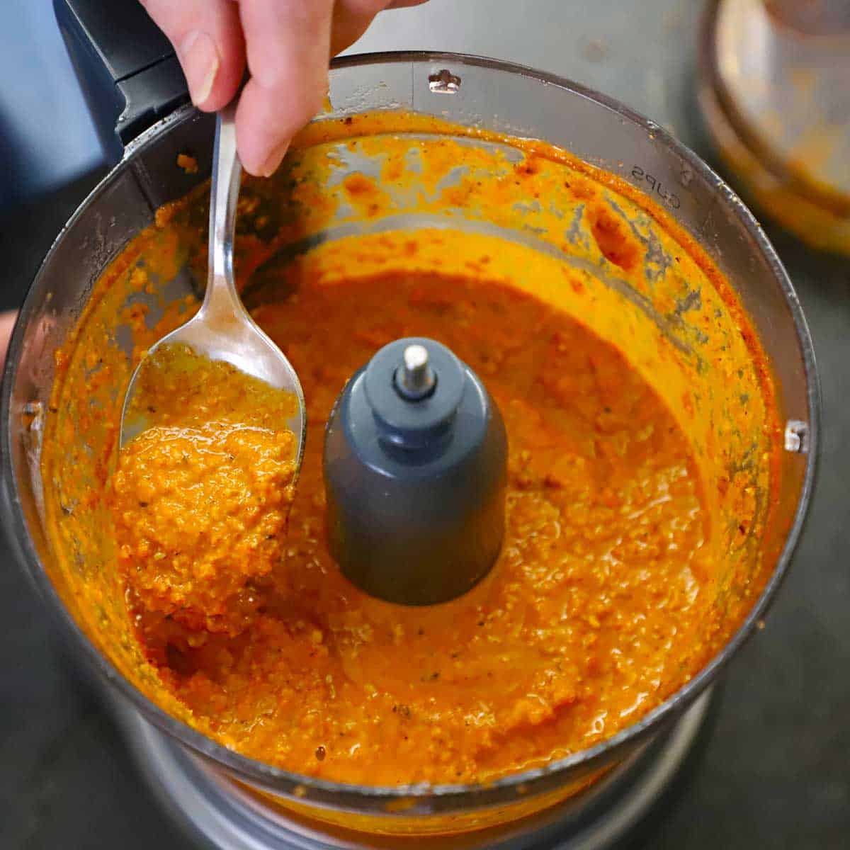 A person using a spoon to hold up a spoonful of smokey romesco sauce that is in the bowl of a food processor.