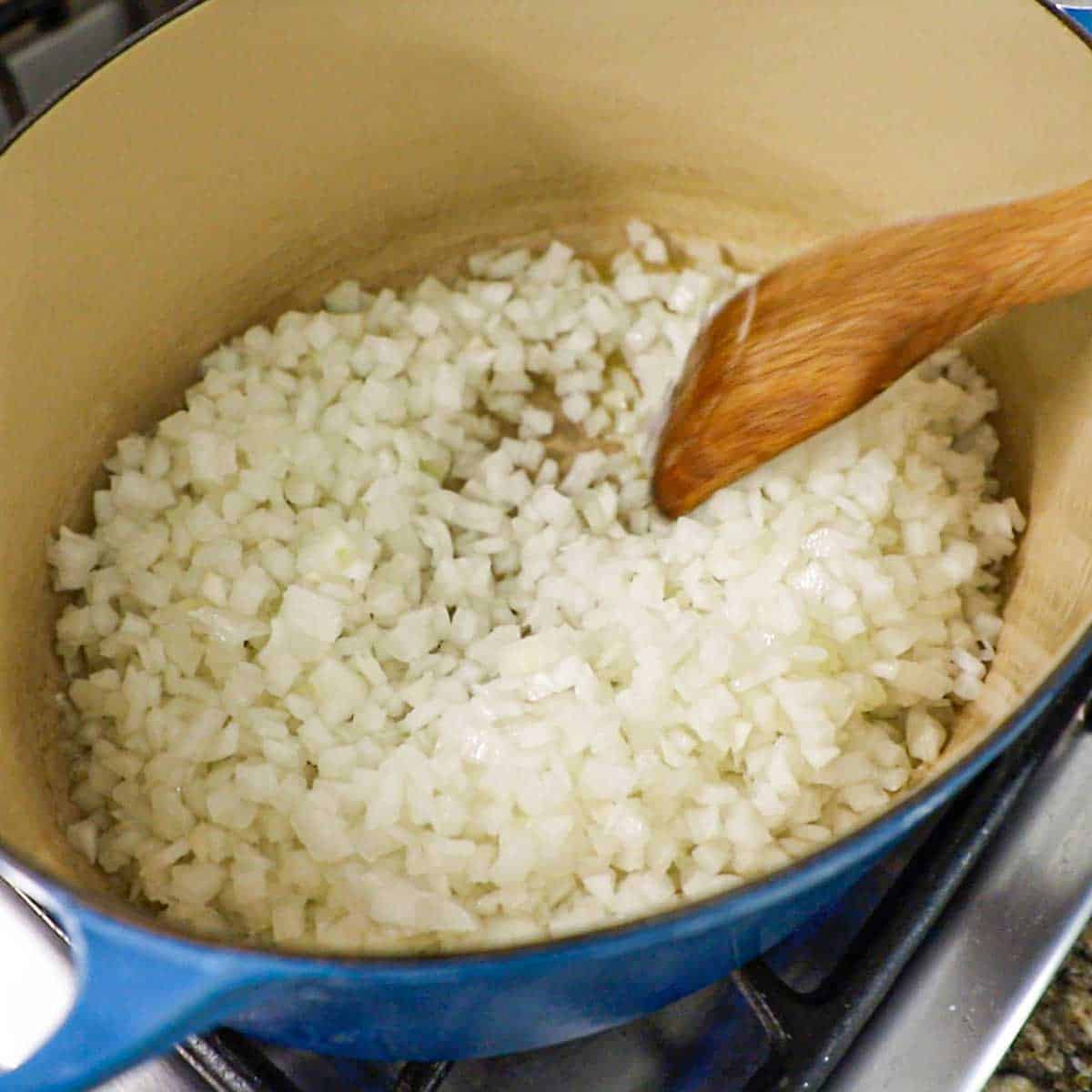 A person using a wooden spatula to stir about 3 cups of chopped onion that are being sautéed in a large oval Dutch oven on a gas stove.