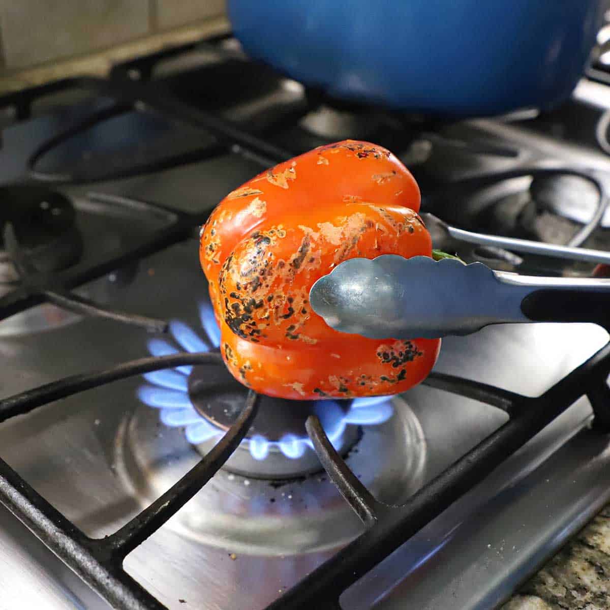 A person using a pair of metal tongs to char a red bell pepper over a gas flame on a gas stove.