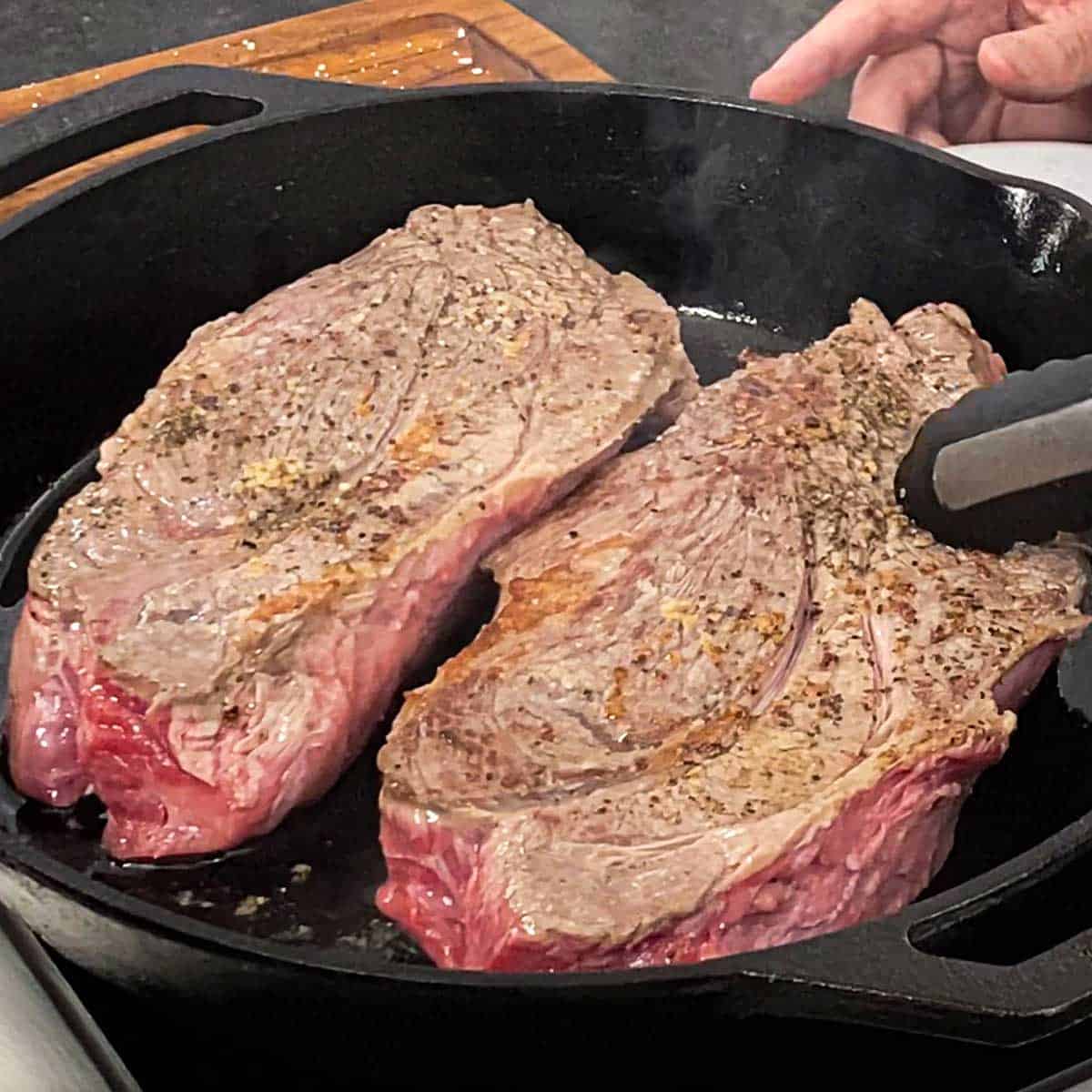 A person using a pair of tongs to flip two seasoned chuck roasts that are being seared in a large cast-iron skillet.