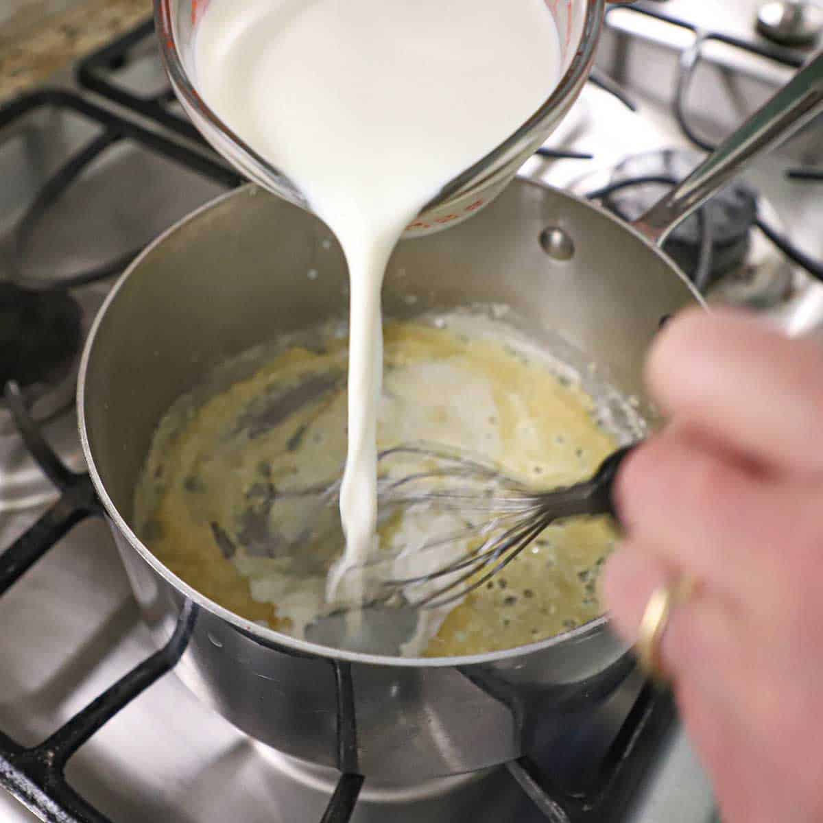 A person using a whisk to combine whole milk being poured from a measuring cup into a saucepan filled with a roux on a gas stove.