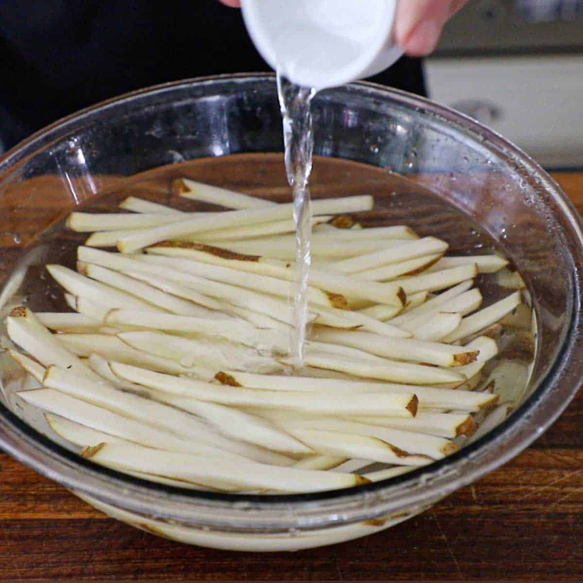 A person pouring distilled white vinegar from a small white bowl into a large glass bowl that is filled with raw strips of potato in the shape of French fries that are submerged in water.