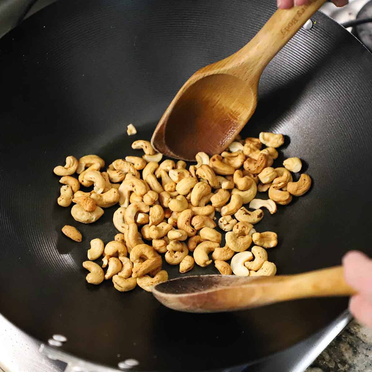 A person using two large wooden spoons to toss raw cashews that are being roasted in a large black wok on a gas stove.