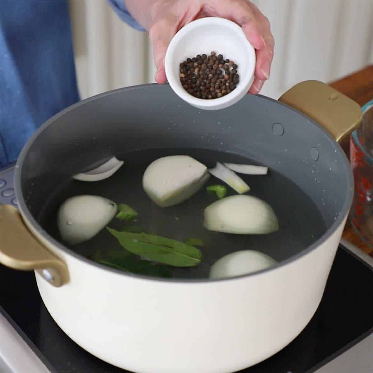 A person adding black peppercorns from a small white bowl into a large pot filled with water, onion quarters, bay leaves, and chopped celery.