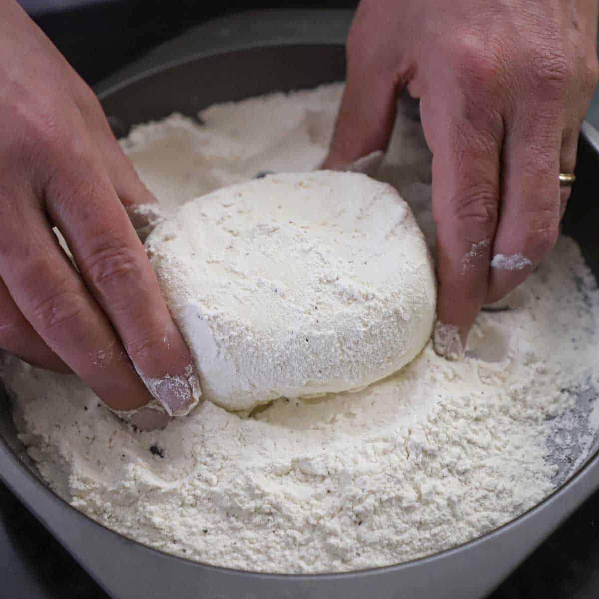 A person using two hands to dredge of large ball of burrata cheese through all-purpose flour that is in a baking pan.