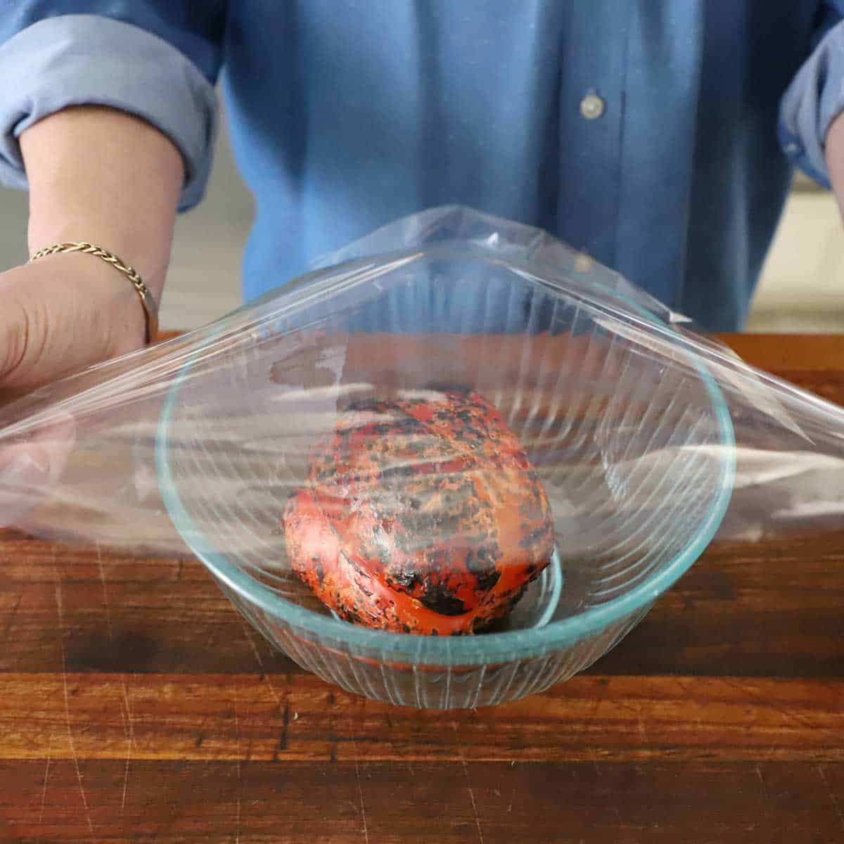 A person placing a piece of plastic wrap over the top of a glass bowl that has a charred red bell pepper in it.