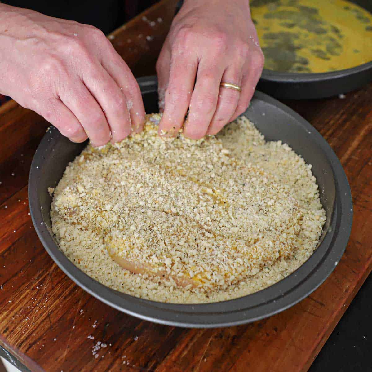 A person dredging a thin chicken cutlet through breadcrumbs in a circular baking pan.