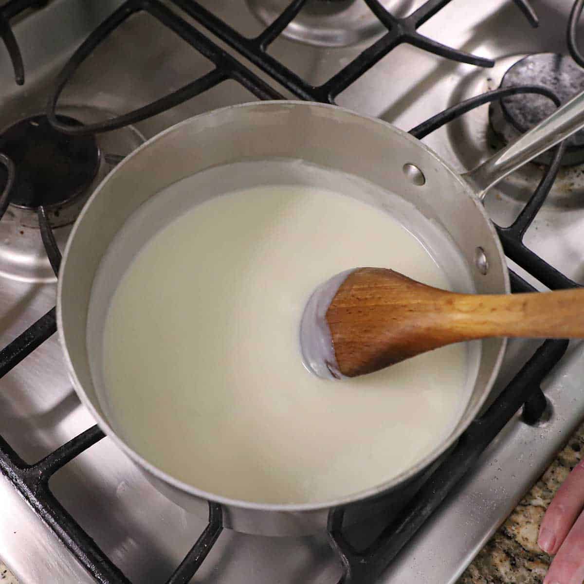 A person using a wooden spoon to stir a bechamel sauce that has slightly thickened in a saucepan on a gas stove.