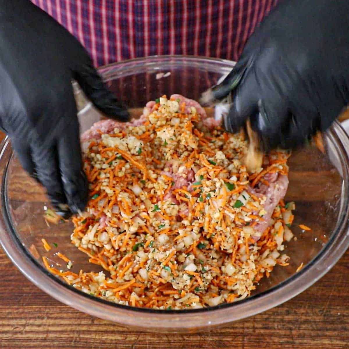 A person wearing black latex gloves mixing the uncooked ingredients for juicy turkey meatloaf in a large glass bowl on wooden cutting board.