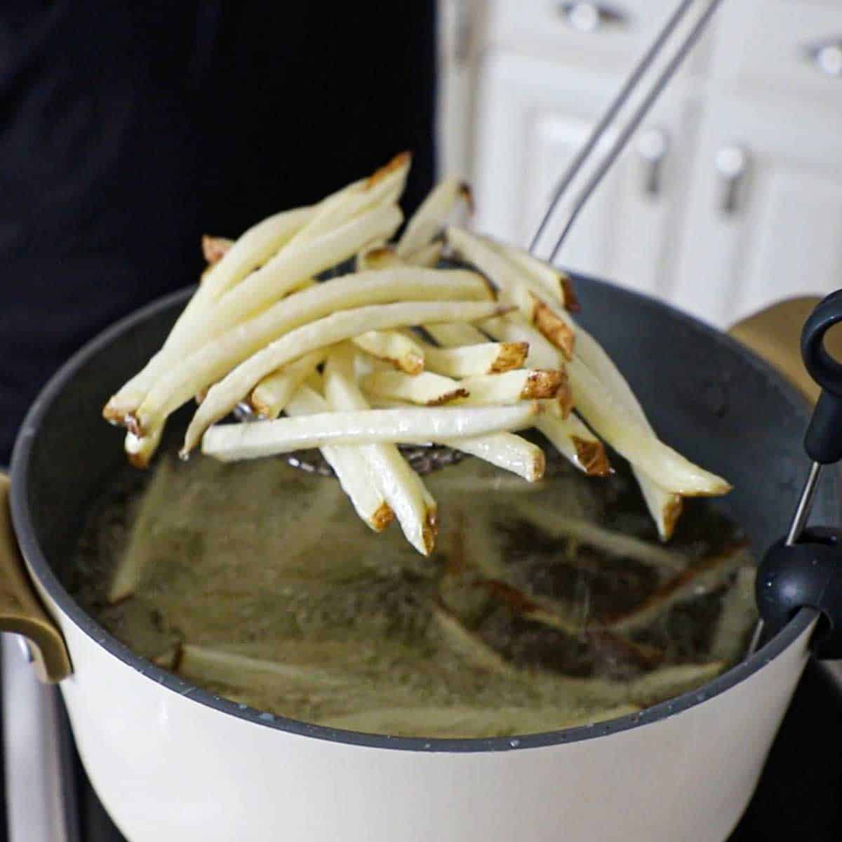 A person using a metal spider to life par-cooked French fries from a large pot of hot oil.
