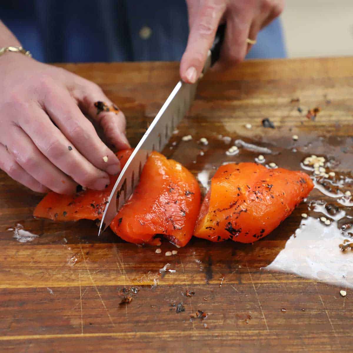 A person using a chef's knife to cut a roasted red bell pepper into large pieces on a wooden cutting board.