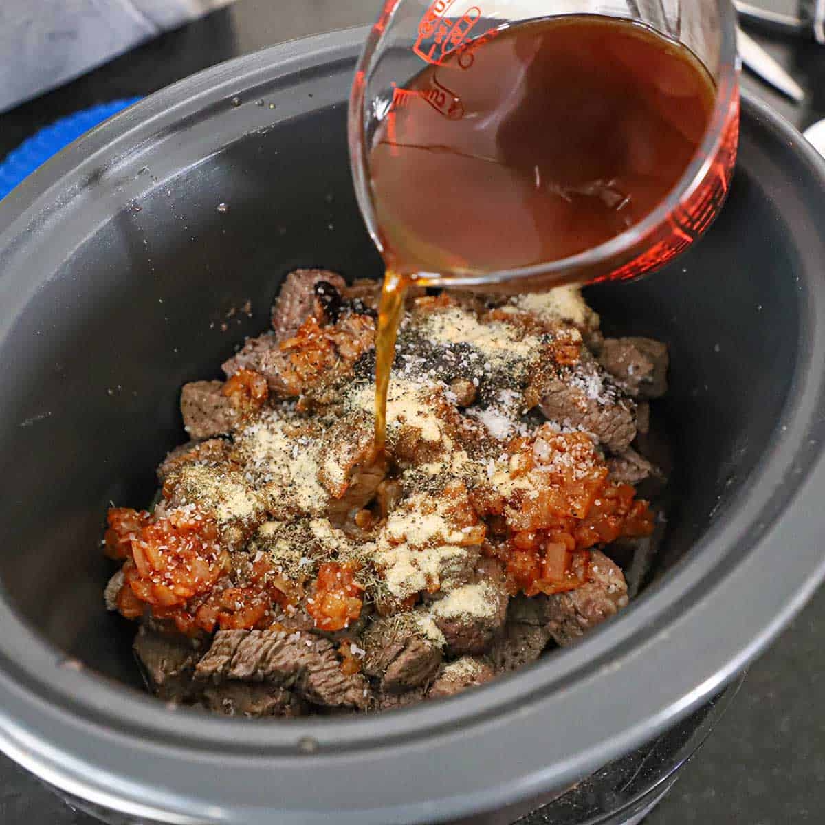 A person pouring beef stock from a 2-cup glass measuring cup into a slow-cooker filled with seared chunks of beef, sautéed onions, and seasonings.