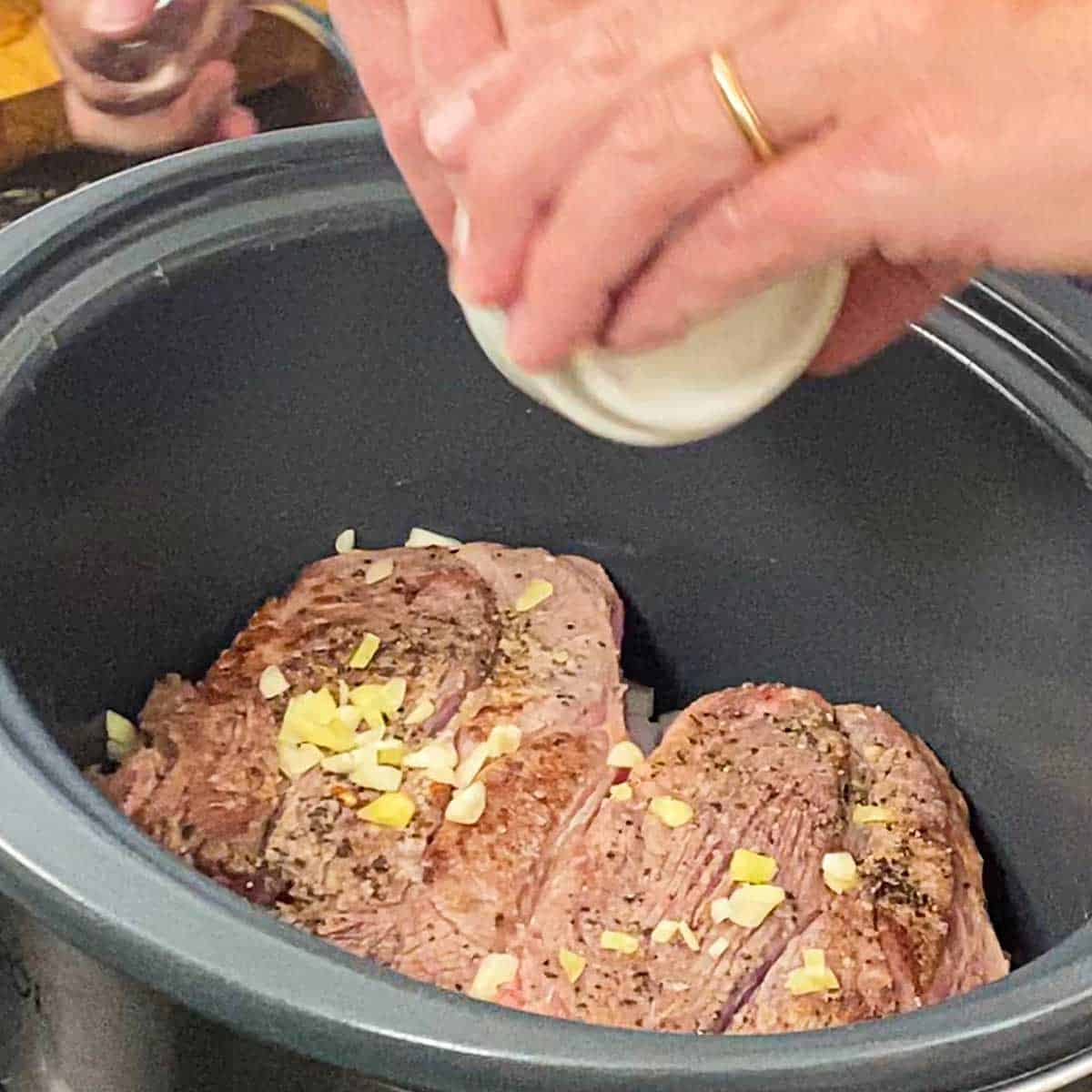 A person adding chopped garlic over the top of two seared seasoned chuck roasts in the bottom of a slow cooker.