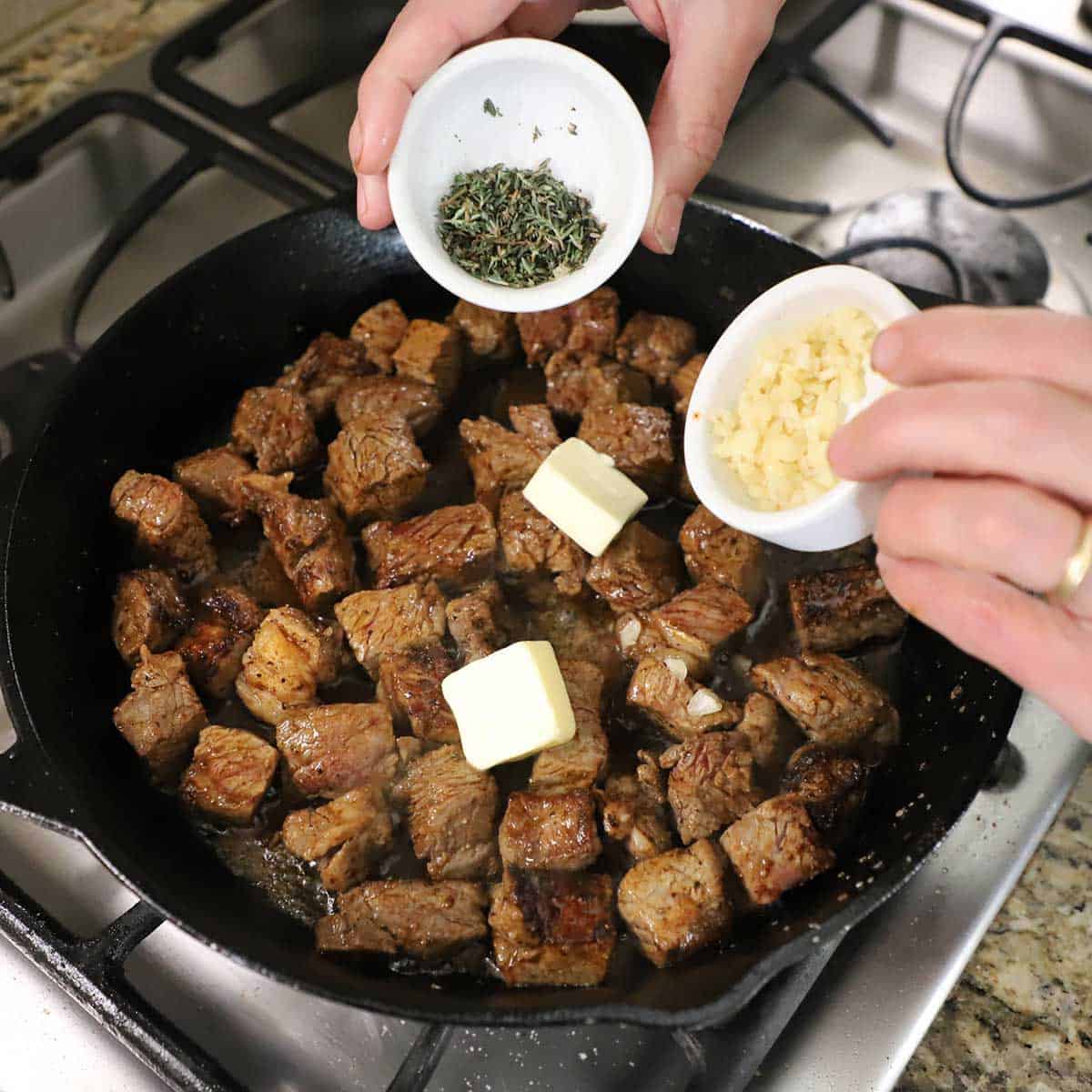 A person dumping chopped fresh thyme from a small white bowl and minced garlic from another small white bowl into a large cast-iron skillet filled with seared steak bitters and two tabs of butter resting on top of the meat.