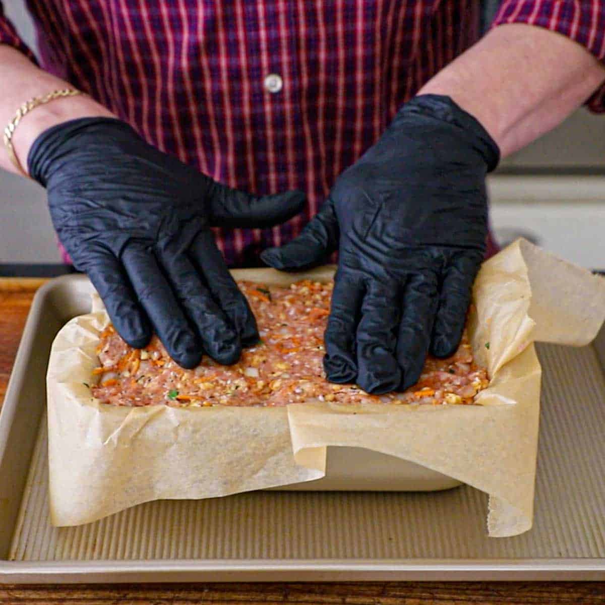 A person wearing black latex gloves gently pressing a juicy turkey meatloaf into a loaf pan that has been lined with parchment paper.