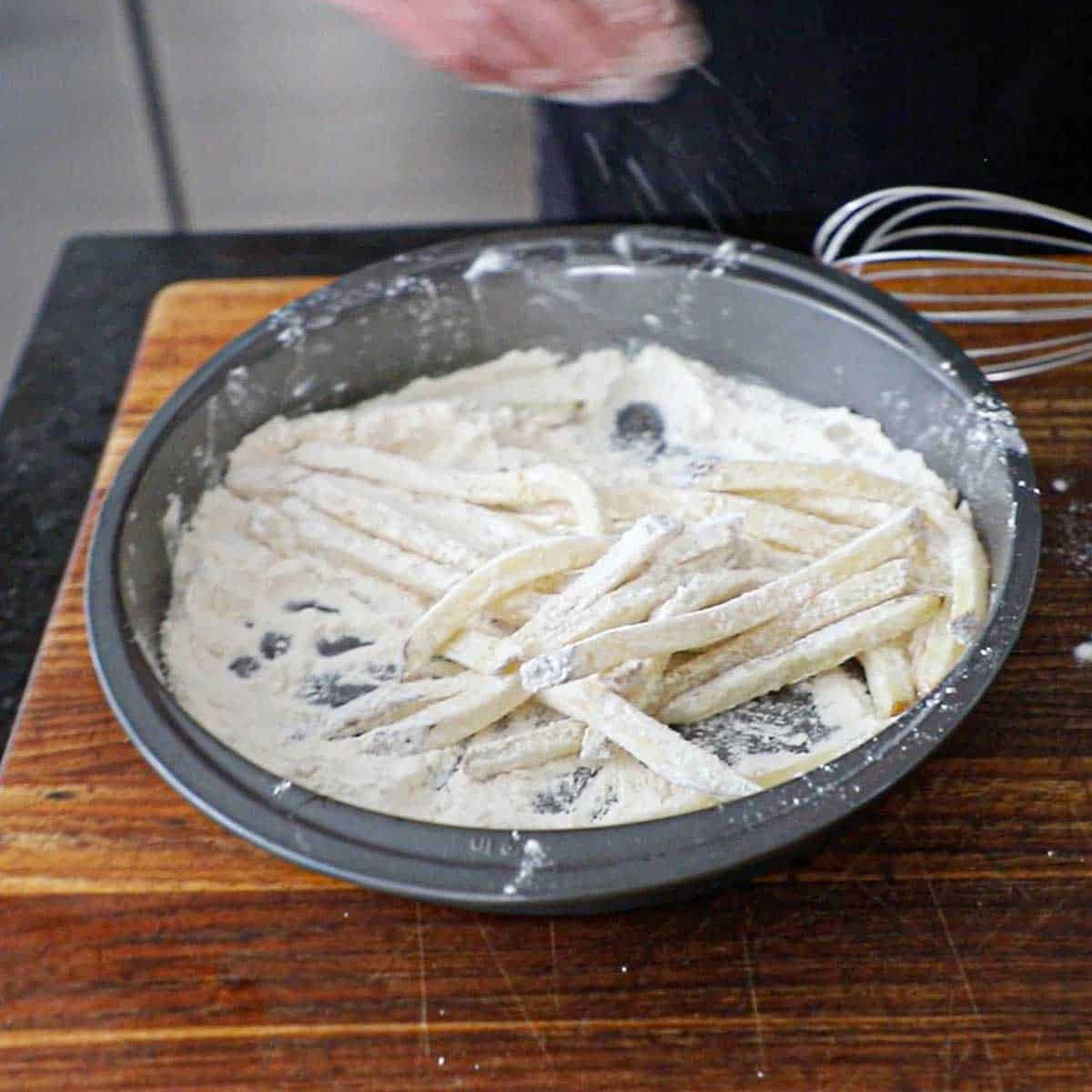A person tossing par-cooked French fries in all-purpose flour in a circular baking pan.