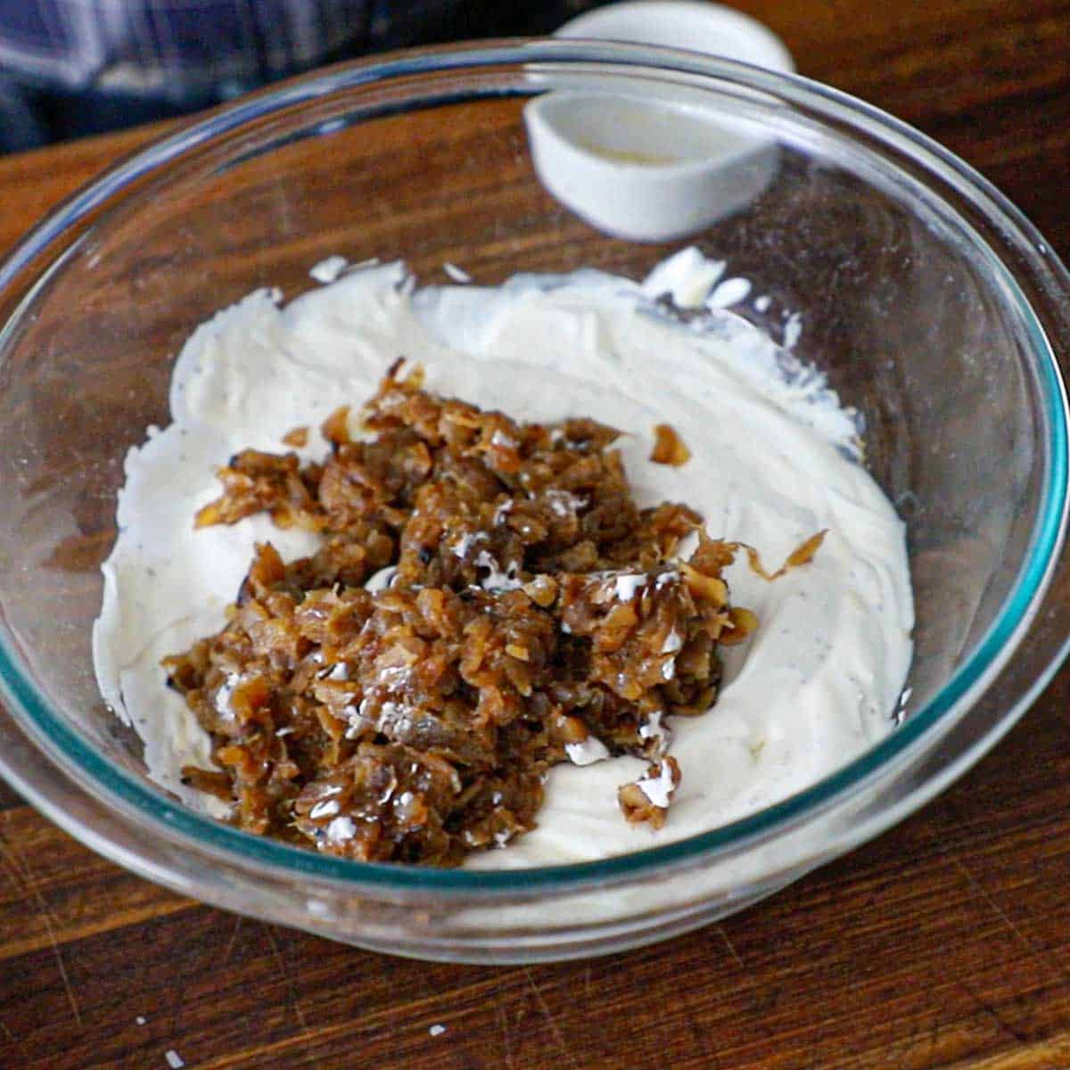 A large glass bowl filled with a seasoned sour cream and mayonnaise mixture with a pile of deeply caramelized onions on top.