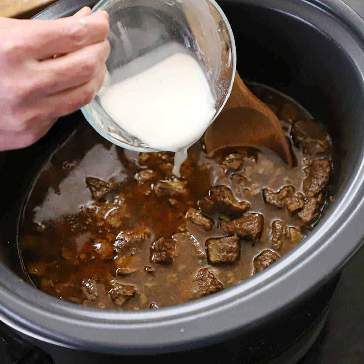 A person pouring a cornstarch slurry from a small glass bowl into a slow cooker that is filled with tender chunks of beef simmering in a dark broth.