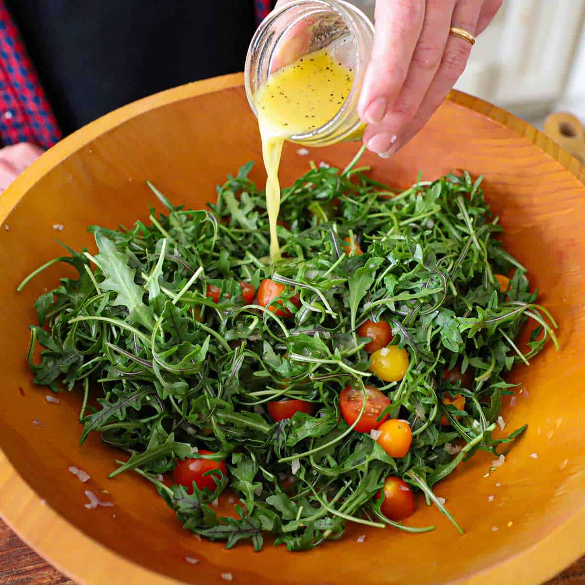A person pouring a Dijon vinaigrette over an arugula and cherry tomato salad in a large wooden salad bowl.