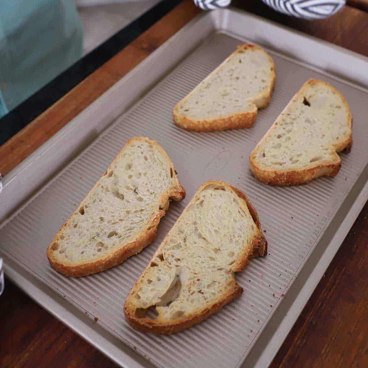 Four slices of toasted sourdough bread on a small sheet pan on a wooden cutting board.
