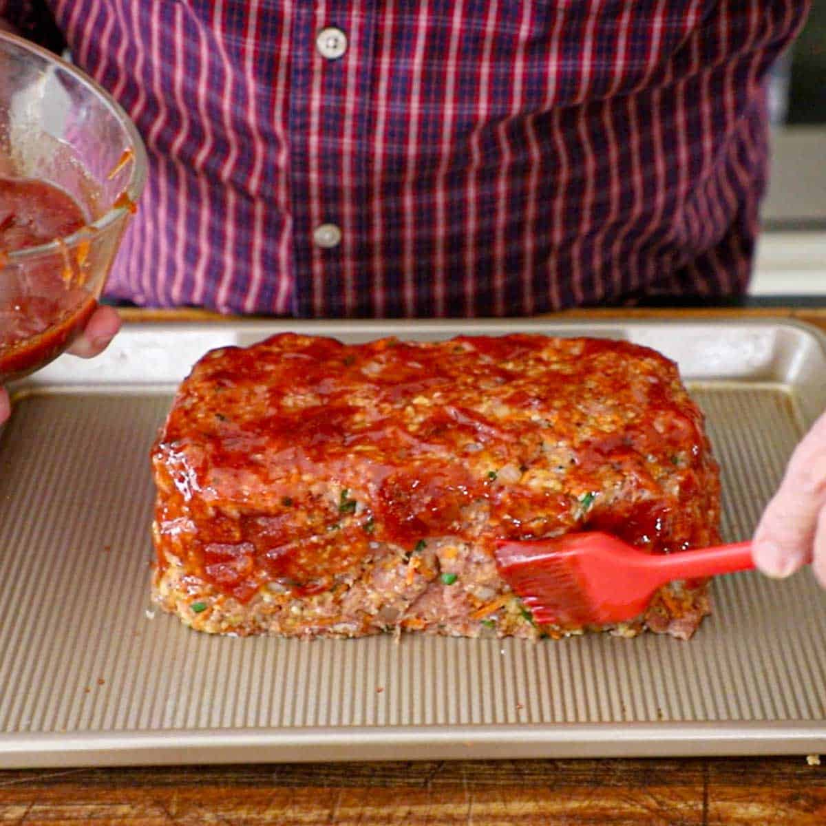A person using a pastry brush to apply a tomato and balsamic glaze onto a loaf-shaped juicy turkey meatloaf on a baking pan.