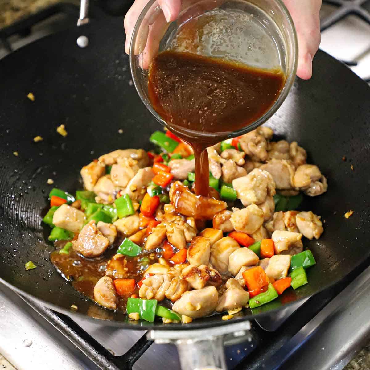 A person pouring a Chinese stir-fry sauce from a small glass bowl into a wok that is filled with cooked chicken pieces and tender pieces of bell peppers.
