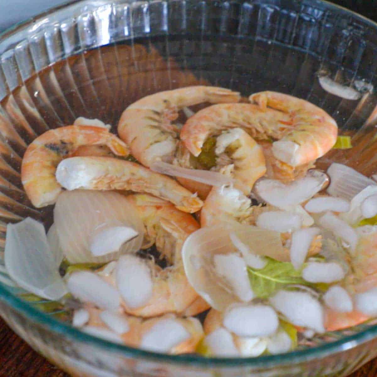 A close-up view of cooked shrimp in an ice bath in a large glass bowl on a cutting board.