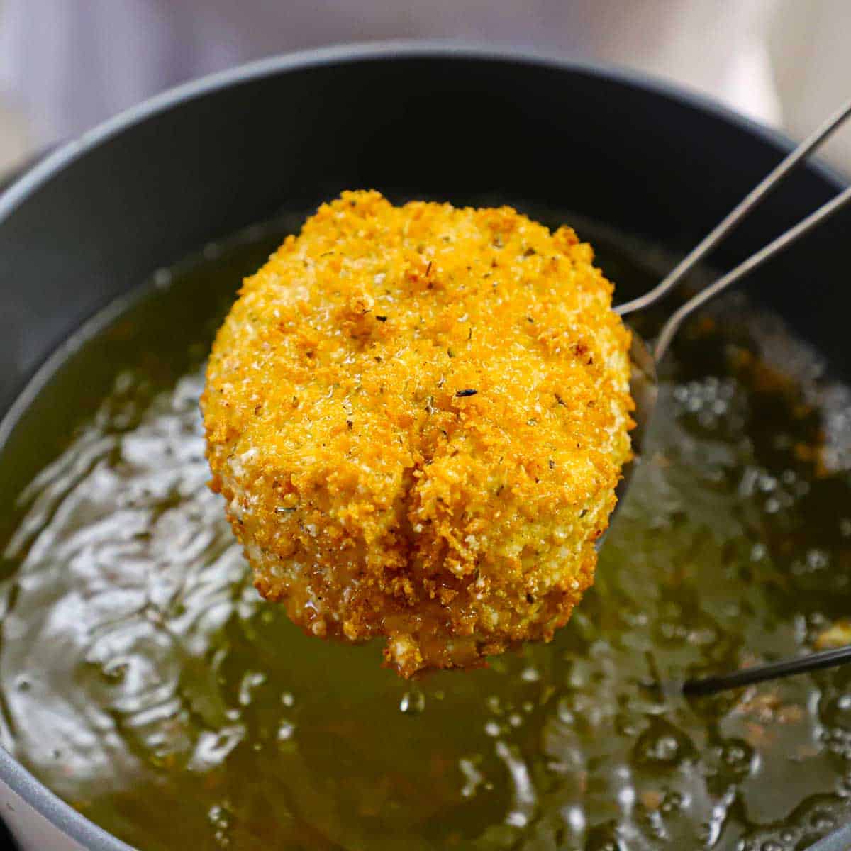 A person using a metal spatula to raise a crispy fried burrata from a pot of hot oil.