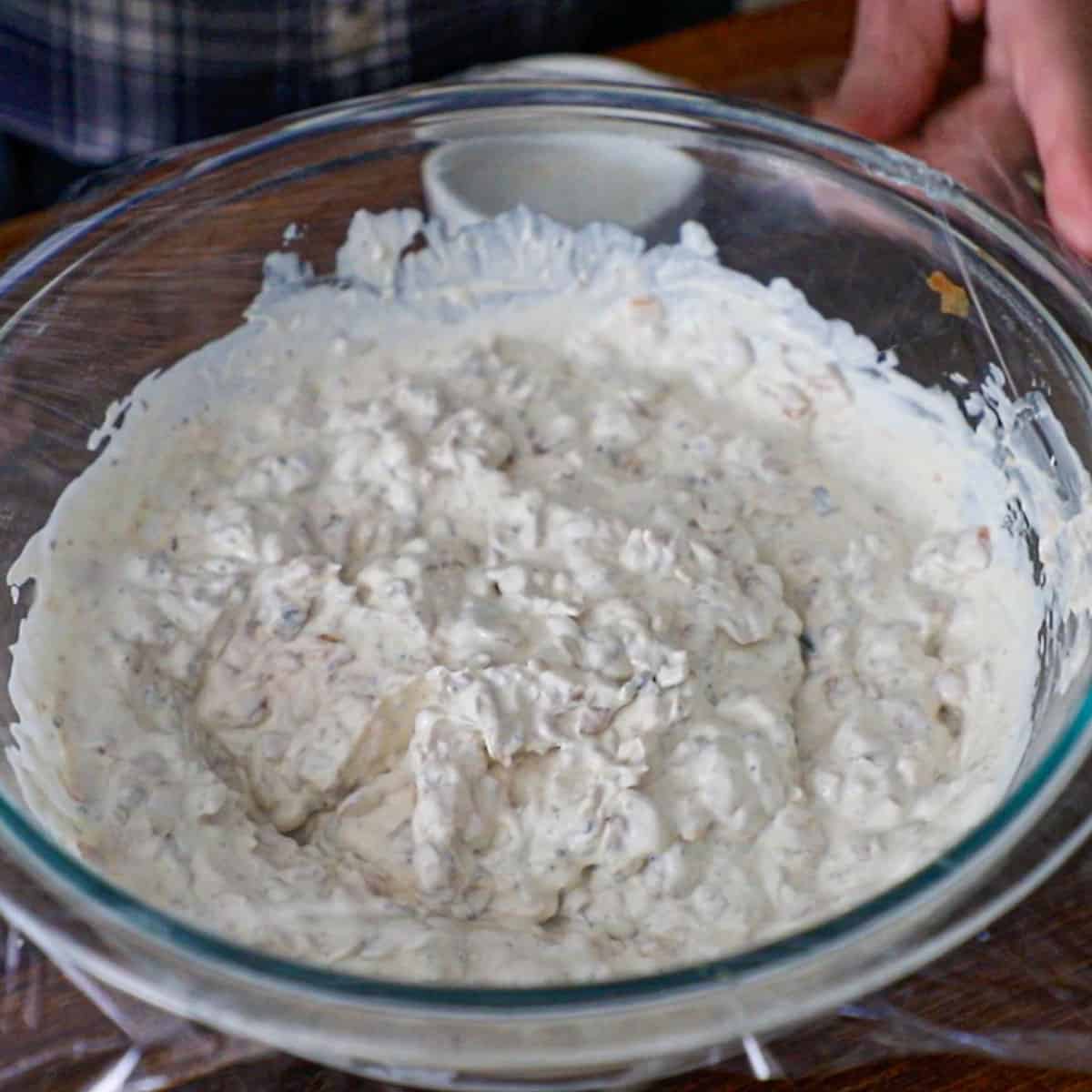 A person placing plastic wrap over the top of a glass bowl that contains freshly made homemade French onion soup.