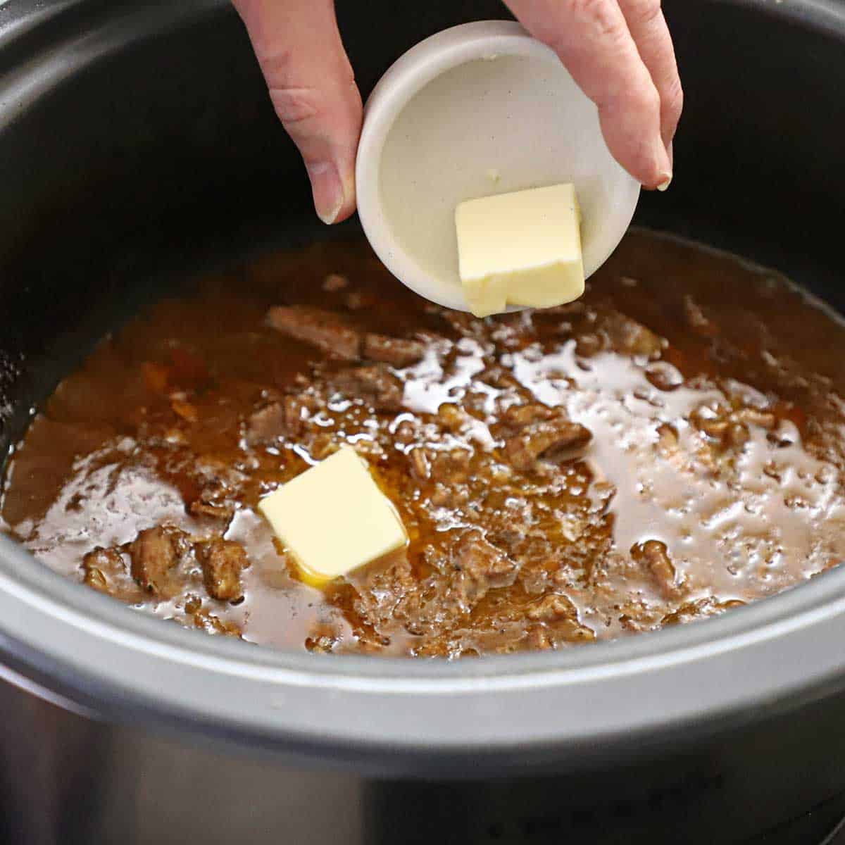 A person dumping two small pads of butter into a slow cooker that is filled with beef tips and gravy.