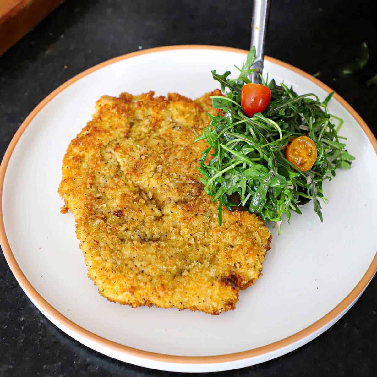 A person using a large spoon to place a mound of an arugula salad on a plate with a crispy chicken Milanese cutlet on it.