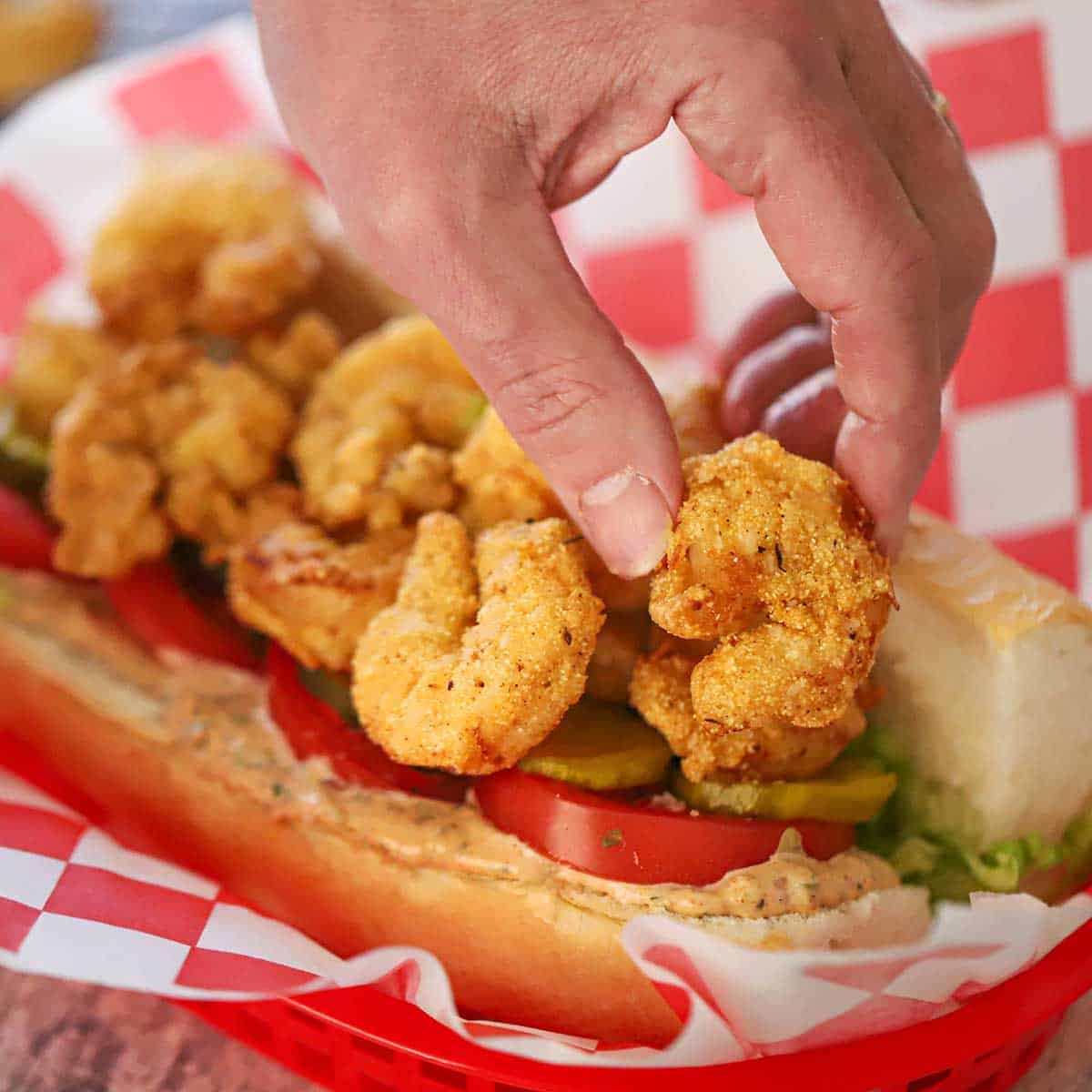 A person placing fried shrimp on top of a French roll that is filled with a layer of Cajun remoulade, then sliced tomatoes, pickles, and shredded lettuce.