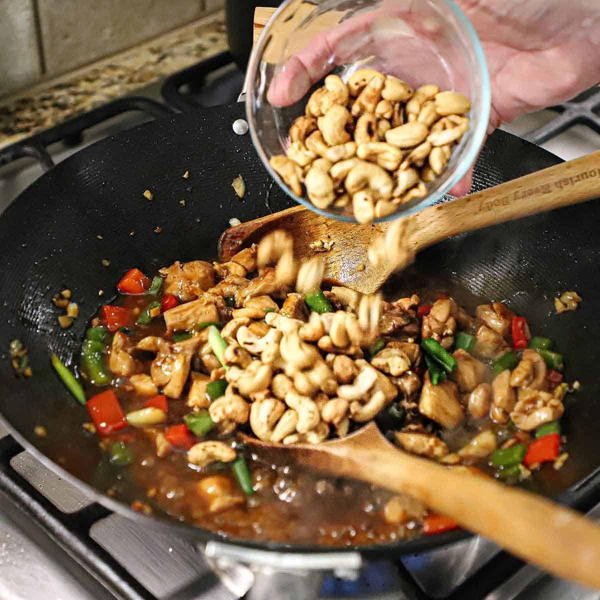 A person transferring roasted cashews from a small glass bowl into a wok that is filled with simmering cashew chicken on a gas stove.