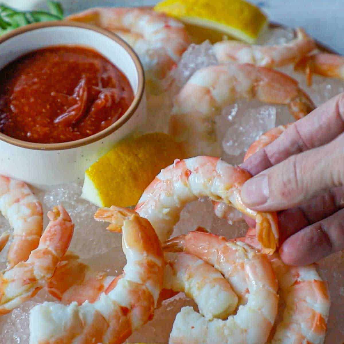 A person placing cooked shrimp onto a plate that is lined with crushed ice and topped with more shrimp, lemon wedges, and a small bowl filled with homemade cocktail sauce.