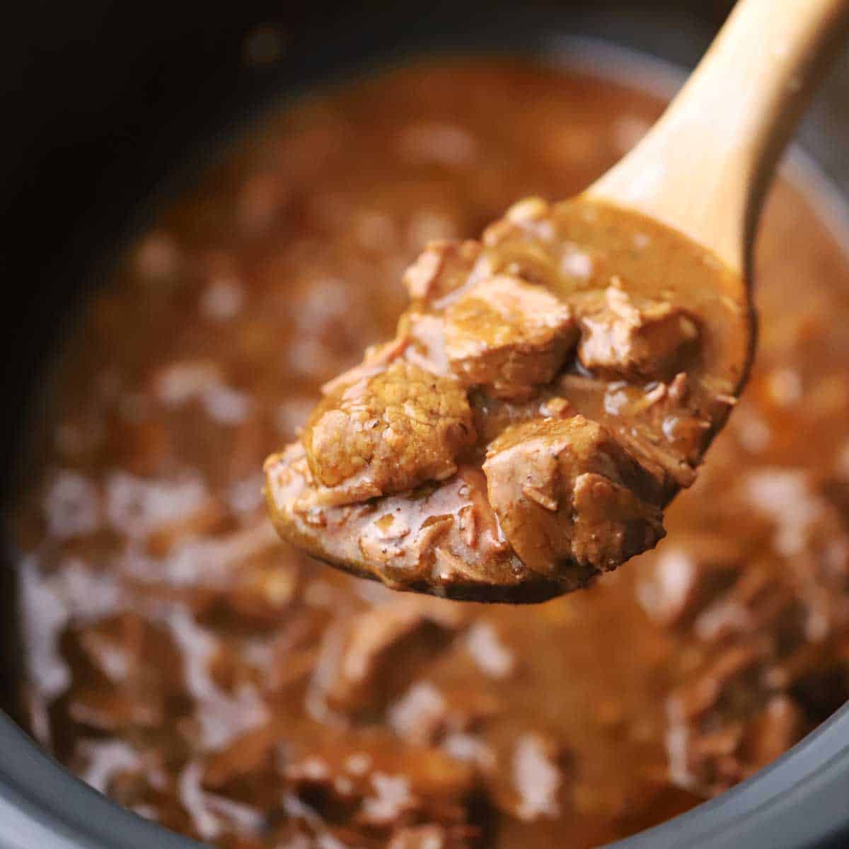 A close-up view of a wooden spoon being used to hold up a serving of slow cooker beef tips and gravy over the slow cooker filled with the same.