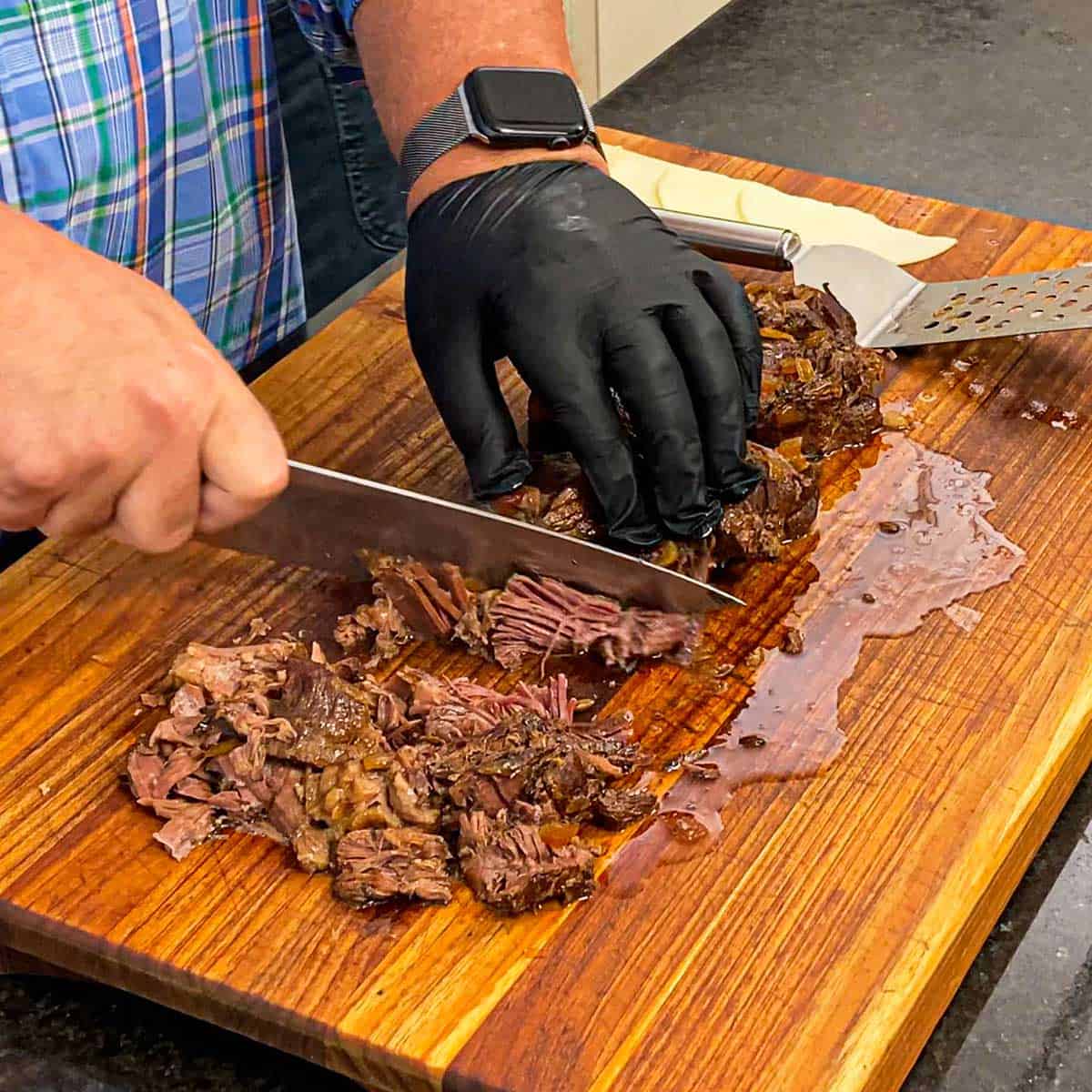 A person using a large knife and his hand to shred and pull apart slow cooked beef for slow cooker French dip sandwiches on a wooden cutting board.