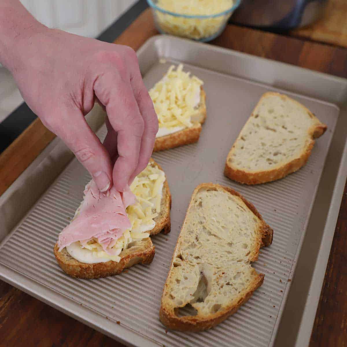 A person placing thinly sliced ham over a slice of toasted bread that has a layer of bechamel sauce and grated Gruyere cheese on it.