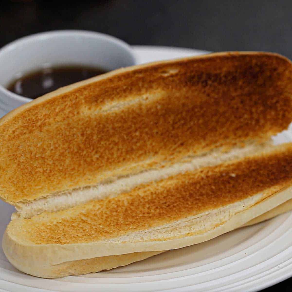 A close-up view of an opened French roll that has been toasted and is resting on a white dinner plate next to a small white bowl filled with au jus sauce.