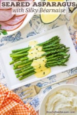 An overhead view of a white rectangle platter filled with a dish of Asparagus with Béarnaise sauce and a large gold serving fork nearby as well as a platter of sliced ham and a bowl of mashed potatoes.