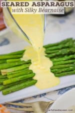A person pouring blender Béarnaise sauce from a glass gravy boat onto a pile of pan-seared asparagus on a white rectangular platter.