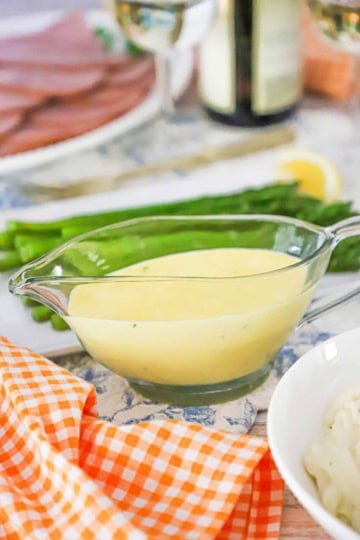A glass gravy boat will with Blender Béarnaise Sauce next to an orange-checkered napkin and a platter filled with pan-seared asparagus.