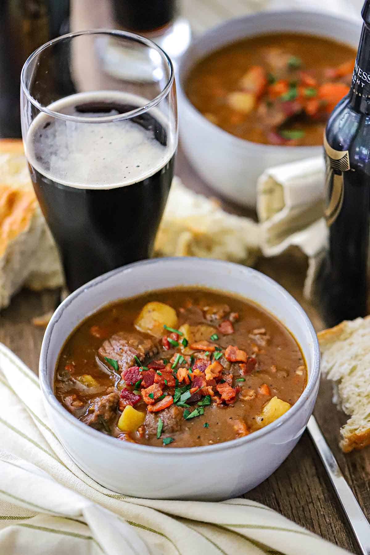 A white soup bowl filled with Classic Guinness Beef Stew topped with crispy bits of bacon and a tall glass of Guinness nearby.